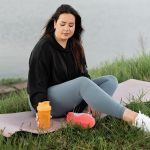 Woman enjoying a peaceful moment by the lake with a yoga mat and water bottle, promoting wellness.